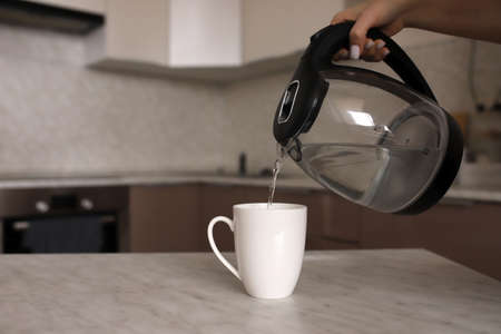 The girl makes tea on the kitchen table. A woman pours boiling water into a white mug on the kitchen table. The girl pours tea into a white mug from a transparent teapot.の写真素材