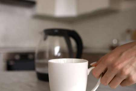 The girl holds with her hand a white mug standing on the table. In the background is a transparent...