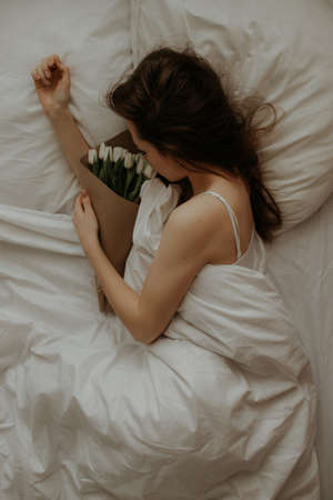 A girl in a white bed with long dark hair lies and hugs a bouquet of white tulips in craft packaging. Girl with bouquets in a white bed. International Women's Day.の写真素材