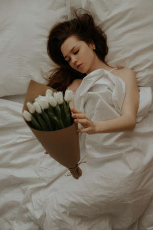 A girl with dark long hair lies on her back in a white bed holding a stylish bouquet of white tulips in craft packaging. The girl in the white bed received a gift for the holiday.の写真素材