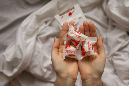 The hands of a young lady are holding a handful of Raffaello sweets on the background of a white bed and a box of chocolates. 03.12.2021 Russia, Altai Territory, Barnaulのeditorial素材