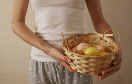 A girl in a white T-shirt against the background of a plain wall stands and holds in her hands a wicker basket with yellow and pink Easter eggs.の写真素材