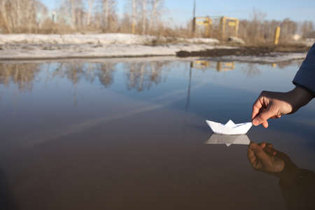 A man's hand launches a paper boat from a checkered notebook sheet in a large puddle in spring. Dirt on the street in spring.の写真素材