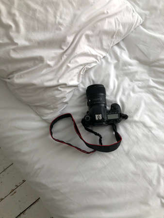 A black SLR camera stands on a white bed, top view. There is a camera on a snow-white bed, a...