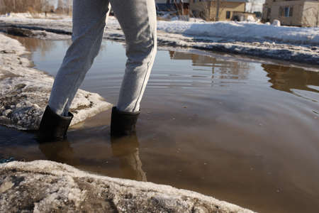 A girl crosses a flooded road in rubber boots. The girl crosses the flooded road with melted snow....