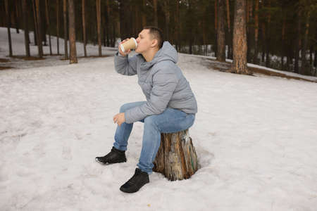 A man sits on a stump in the forest in a gray jacket and drinks coffee. a young man in the forest in early spring sits on a stump and drinks coffee.の写真素材