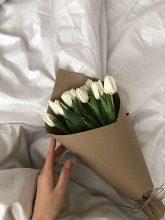 A bouquet of white tulips lies on a white bed. A bouquet of tulips is packed in craft paper and lies on the bed, vertical photo.の写真素材