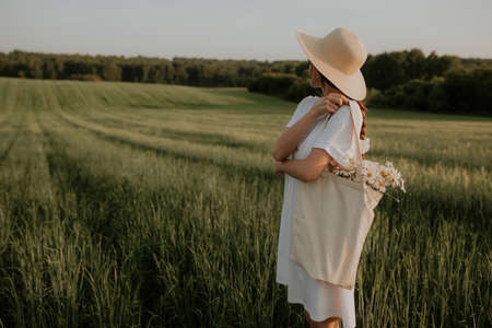 A girl in a white dress stands in a green field looking into the distance. A young woman stands in a...