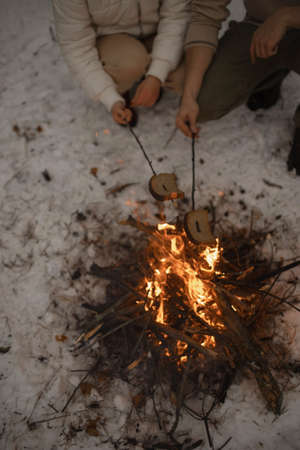 A young couple roast bread on a campfire in a snowy forest. Young couple fry bread outdoors in winter over an open fire.の写真素材