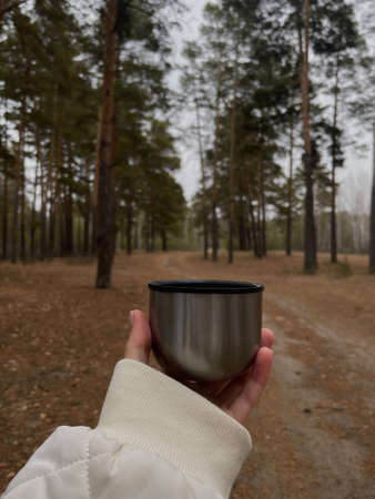A girl's hand holds a glass from a thermos in the autumn forest. The girl's hand holds a mug with...