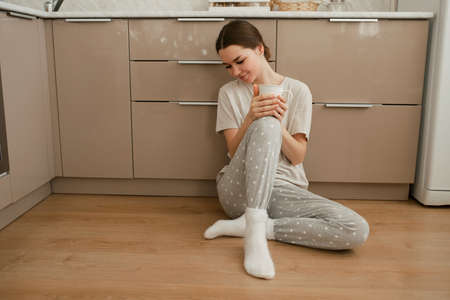 A happy young girl sits on the kitchen floor and drinks tea. The girl is drinking tea while sitting on the floor at home.の写真素材