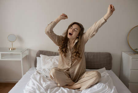 A young girl in beige pajamas is hunched over in bed. The girl yawns in the morning sitting in bed. selective focusの写真素材