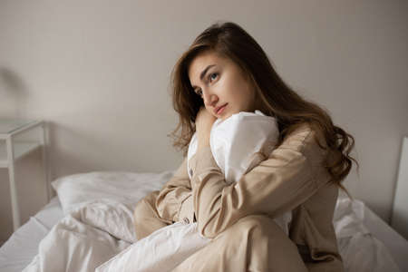 A sad girl sits in bed hugging a white blanket with a pensive face. Thoughtful girl in pajamas sits in bed.の写真素材