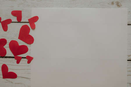 Red paper hearts are scattered on a white wooden background. A sheet of text on the table next to red hearts.の写真素材