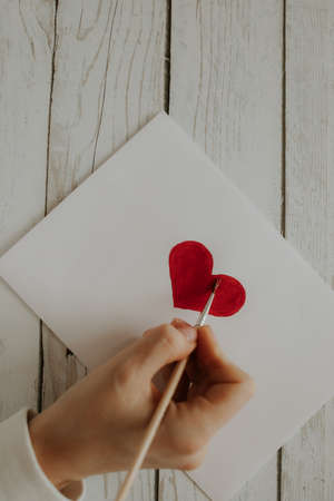 The red heart is painted on white paper. The girl draws a red heart with a brush on white paper....