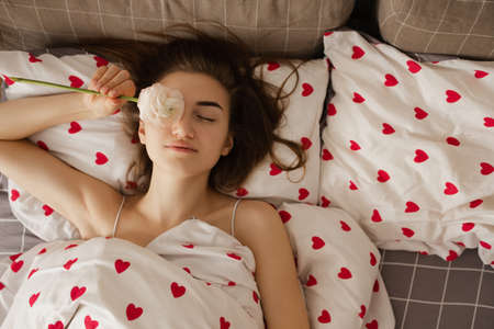 The girl lies in bed. The girl lies in bed with red hearts and holds a flower in her hand.