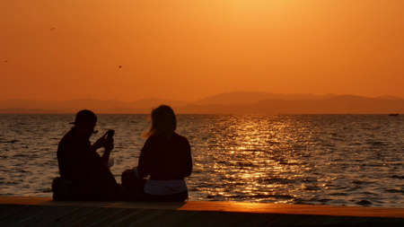 Silhouette of a young couple taking a selfie at sunset, with a sea and sunset in the backgroundの写真素材