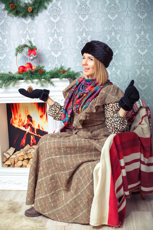 attractive blonde woman in a brown dress and a black fur hat near Christmas tree and fireplaceの写真素材