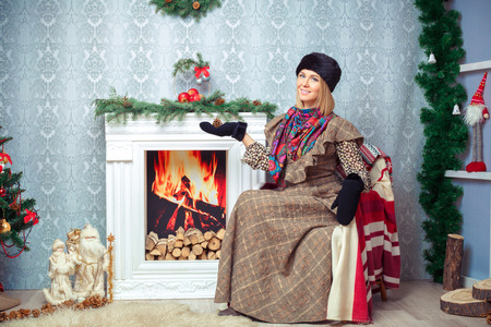 attractive blonde woman in a brown dress and a black fur hat near Christmas tree and fireplaceの写真素材
