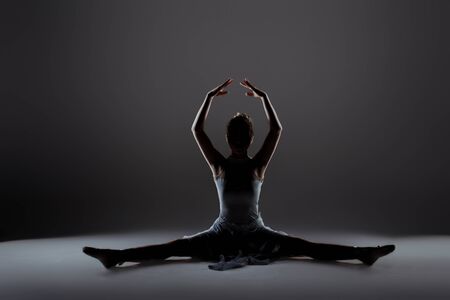young and beautiful dancer girl doing stretching exercises. This is to warm up before exercise to increase the elasticity of muscles. ballerina warming-up. silhouette in studioの写真素材