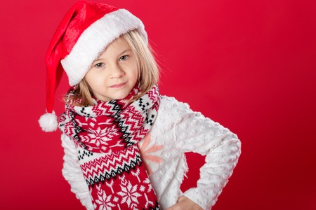 portrait of a little smiling blonde girl in santa hat and scarf on red backgroundの写真素材