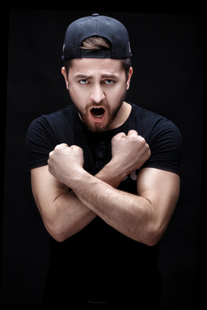 angry young man in the black shirt and cap screaming on black background.の写真素材