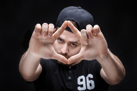 portrait of young man with dark hair in shirt and hat on black background. Mans hands making frameの写真素材