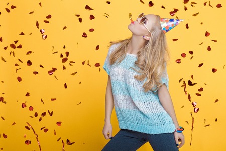 portrait of funny blond woman in birthday hat and red confetti on yellow background. Celebration and...