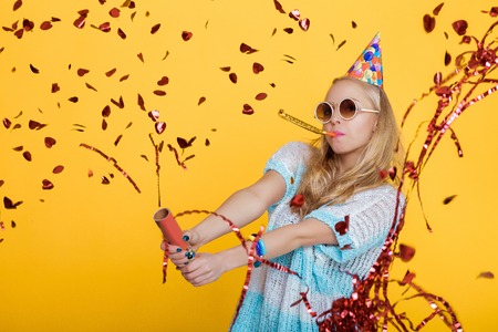 portrait of funny blond woman in birthday hat and red confetti on yellow background. Celebration and party.の写真素材