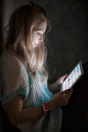 portrait of young blonde woman looks and reads on tablet and drinking coffee at night. girl uses the tabletの写真素材