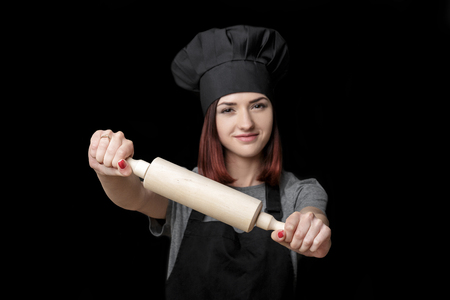 Young attractive woman chef in black uniform holds rolling pin  on  black background. Focus on rolling pinの写真素材