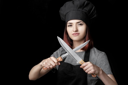 Young attractive woman chef in black uniform holds Two knives on black background. Focus on knivesの写真素材
