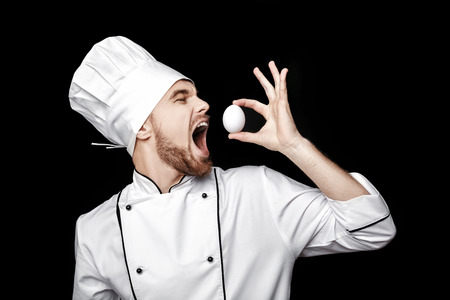 Young bearded man chef In white uniform holds  egg on black backgroundの写真素材
