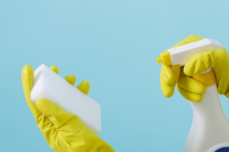 Hand in yellow glove holds spray bottle of liquid detergent and sponge on blue background. cleaningの写真素材