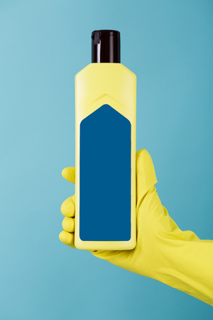 Hand in rubber yellow glove holds a bottle of liquid detergent on blue background. cleaningの写真素材