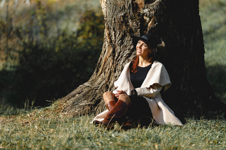 woman in a beige coat and black hat sitting under treeの写真素材