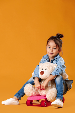 Stylish little child girl with skateboard in denim on orange backgroundの写真素材