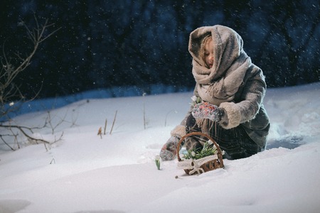 cute little child girl collects in basket first flowers under snow in winter evening. fantastic night. frame as illustration to the fairy tale 12 monthsの写真素材