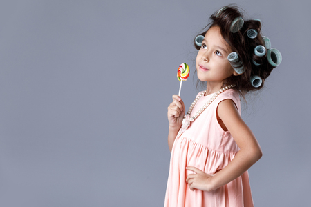 Funny little child girl in pink dress and hair curlers holding lollipop on gray background.の写真素材