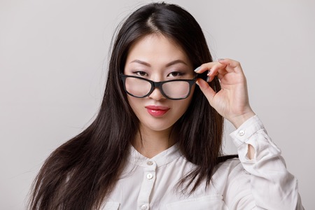 portrait of young smiling asian woman in glasses and white shirt looking to camera on gray background.の写真素材