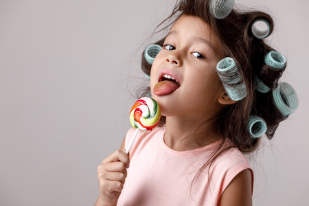 Funny little child girl in pink dress and hair curlers eating lollipop on gray background.の写真素材