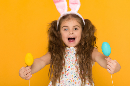 Portrait of surprised little child girl with Easter bunny ears holding colorful eggs on yellow background. Happy easterの写真素材