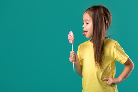 Beautiful cute little child girl eating sweet candy lollipop isolated on blue backgroundの写真素材