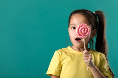 Beautiful surprised little child girl with sweet candy lollipop isolated on blue backgroundの写真素材