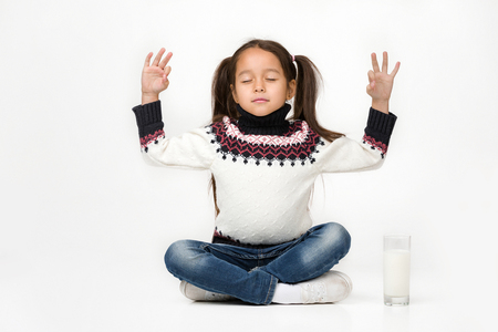 Portrait of cute little child girl sitting in lotus position and a glass of fresh milk is standing nearbyの写真素材
