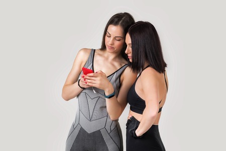 two beautiful athletic woman friends using app on mobile phone, looking at screen of smart phone on break between exercisesの写真素材