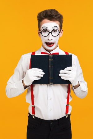 portrait of surprised mime man artist reading book isolated on yellow backgroundの写真素材