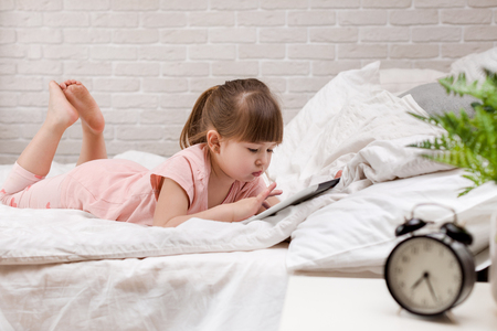 cute little child girl lies in bed uses digital tablet. child playing on tablet pc.の写真素材