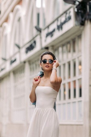 portrait of attractive woman in sunglasses and white dress walks through the city streets on a sunny day. girl in the old townの写真素材