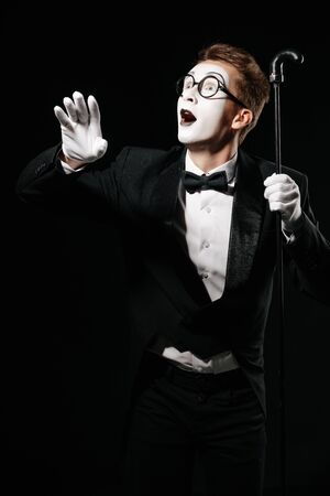 surprised mime man in tuxedo and glasses posing with walking stick on black backgroundの写真素材